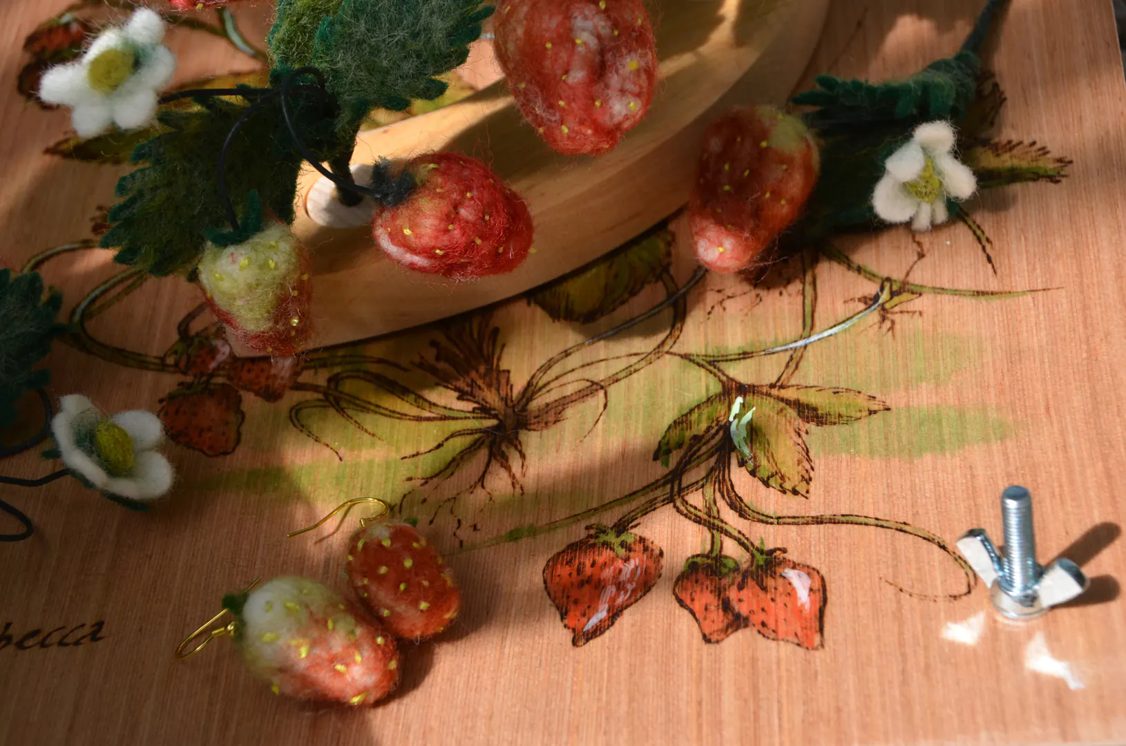 Een verzameling van handgemaakte aardbei-producten: een houten bloemenpers, vilten oorhangers en jaarringstekers. Een tastbare weergave van de zomer, gemaakt van natuurlijke materialen.