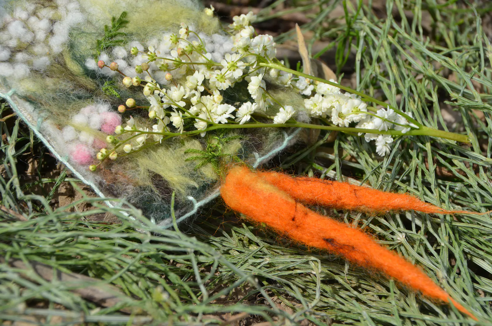 Handgemaakt vilten werkje van wilde peen, buiten de kaders. Versierd met verse filipendula knolspirea uit eigen seizoenstuin voor een tastbare natuurweergave.